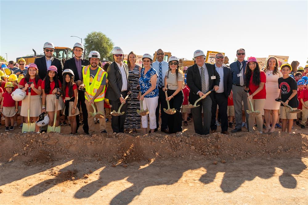 People dressed in hard hats and safety vests use shovels to dig into the ground